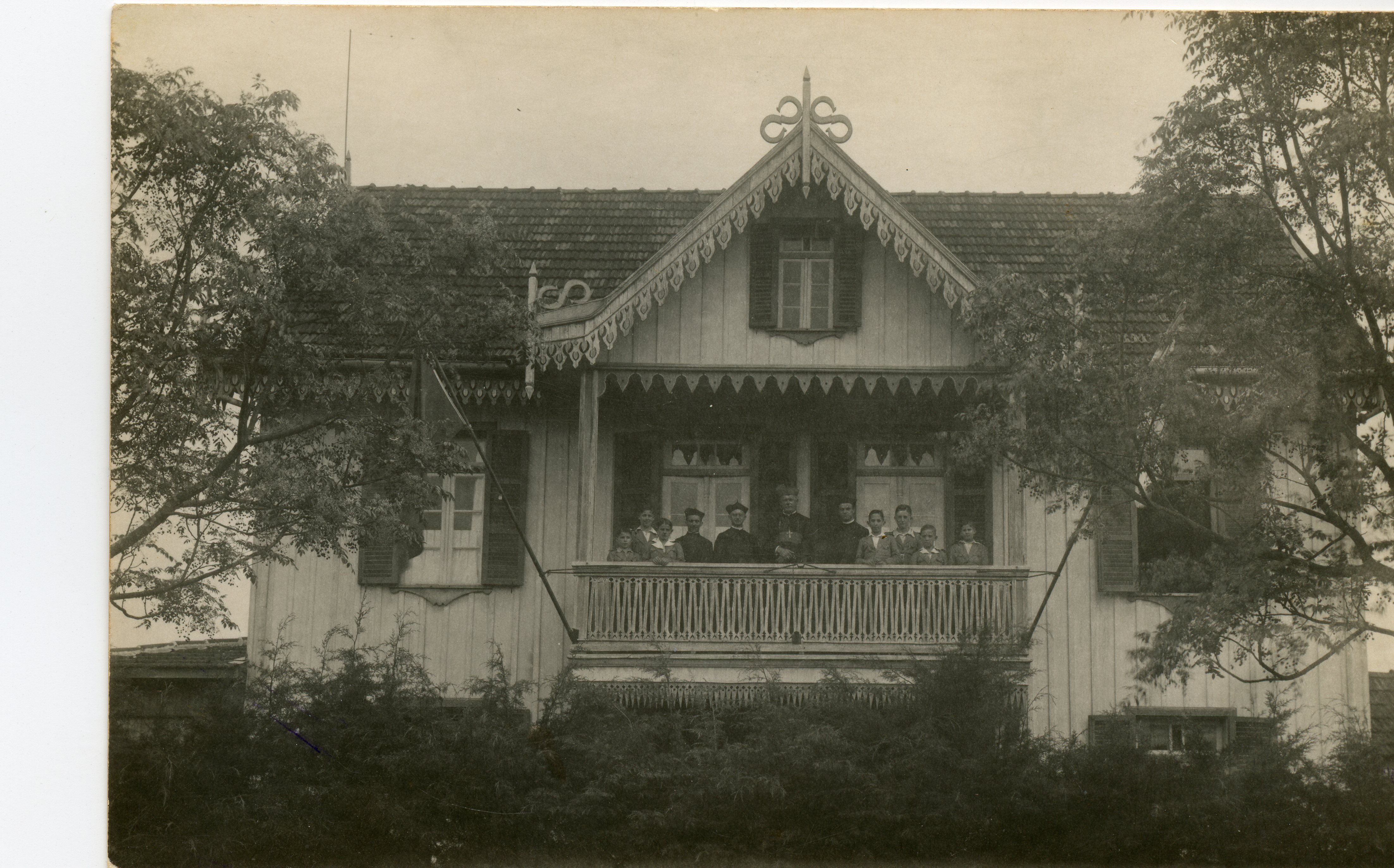 Fotografia em preto e branco de uma residência de madeira com arquitetura típica de influência europeia, possivelmente de colonização alemã ou italiana no sul do Brasil. Um grupo de pessoas, incluindo figuras que parecem ser religiosos, está posicionado na varanda do andar superior da casa, cercada por vegetação.