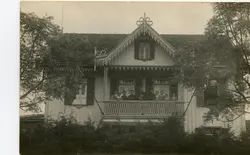 Fotografia em preto e branco de uma residência de madeira com arquitetura típica de influência europeia, possivelmente de colonização alemã ou italiana no sul do Brasil. Um grupo de pessoas, incluindo figuras que parecem ser religiosos, está posicionado na varanda do andar superior da casa, cercada por vegetação.