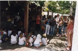 Uma cena ao ar livre sob uma parreira, onde um grupo de jovens músicos toca instrumentos de sopro para uma plateia de crianças sentadas no chão. O ambiente sugere uma atividade cultural ou educacional em uma região de colonização italiana, possivelmente em Videira, com vestimentas típicas do início dos anos 2000.