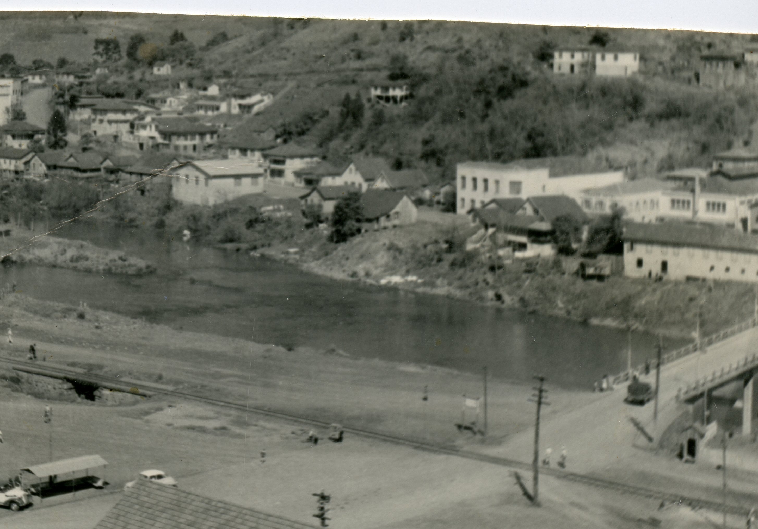 Fotografia panorâmica em preto e branco de uma pequena cidade brasileira, possivelmente Videira, mostrando um rio que corta o centro urbano. A imagem exibe construções de época, uma ponte e uma linha férrea, sugerindo um cenário de desenvolvimento urbano no início ou meados do século XX.