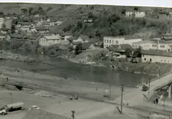 Fotografia panorâmica em preto e branco de uma pequena cidade brasileira, possivelmente Videira, mostrando um rio que corta o centro urbano. A imagem exibe construções de época, uma ponte e uma linha férrea, sugerindo um cenário de desenvolvimento urbano no início ou meados do século XX.