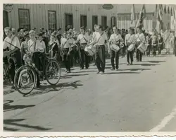 Fotografia em preto e branco de um desfile cívico em uma rua de terra, provavelmente em uma pequena cidade brasileira como Videira. Jovens uniformizados com camisas brancas e faixas a tiracolo marcham tocando tambores, acompanhados por um ciclista, em frente a construções de madeira típicas da colonização sulista, sugerindo um evento comemorativo de meados do século XX.