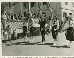 Uma fotografia em preto e branco que retrata um desfile cívico ou evento comunitário, provavelmente em uma pequena cidade brasileira. Jovens estudantes uniformizadas e um homem adulto carregam bandeiras, enquanto uma multidão observa de uma estrutura de madeira elevada e das calçadas, sugerindo um ambiente de celebração local de meados do século XX.