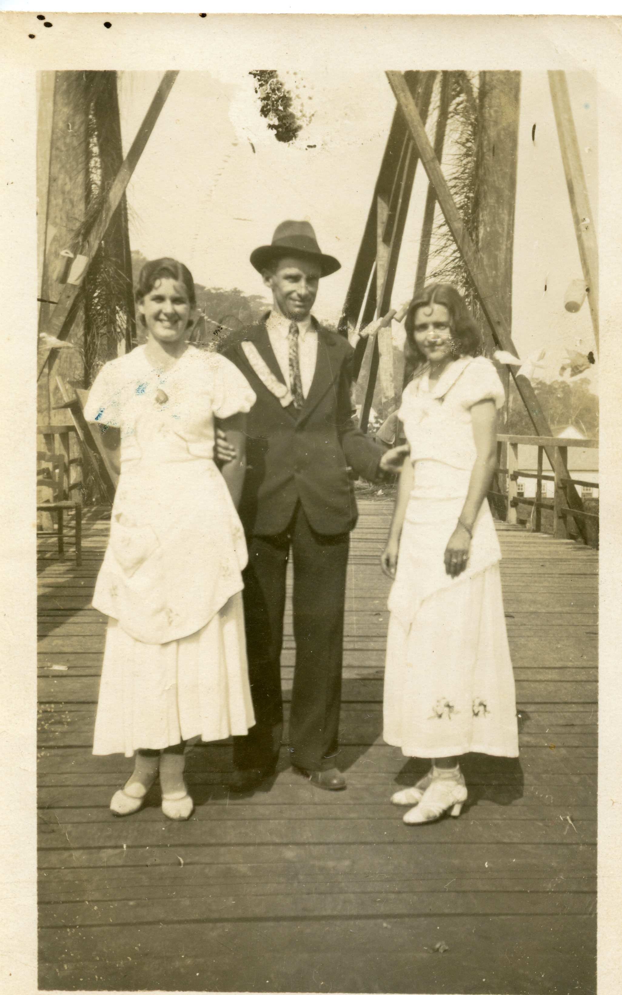 Uma fotografia em sépia retratando um homem de terno e chapéu posando entre duas mulheres vestidas com trajes claros, possivelmente em uma ponte de madeira com estrutura metálica. A vestimenta e o estilo da imagem sugerem um contexto rural ou de pequena cidade brasileira nas décadas de 1930 ou 1940.