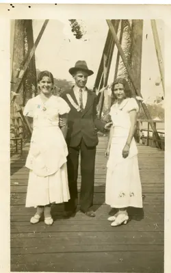 Uma fotografia em sépia retratando um homem de terno e chapéu posando entre duas mulheres vestidas com trajes claros, possivelmente em uma ponte de madeira com estrutura metálica. A vestimenta e o estilo da imagem sugerem um contexto rural ou de pequena cidade brasileira nas décadas de 1930 ou 1940.