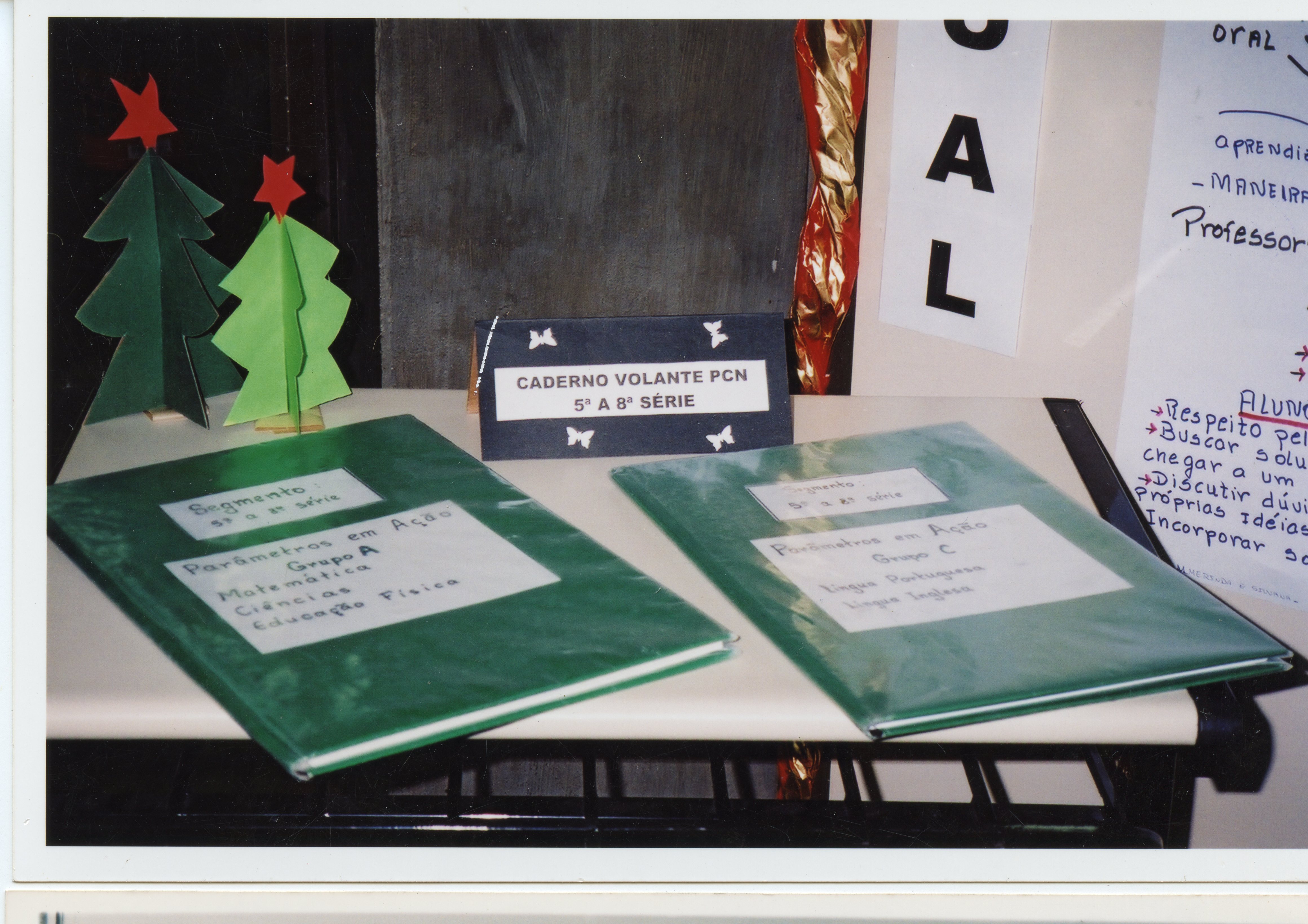 A imagem mostra uma exposição escolar sobre os Parâmetros Curriculares Nacionais (PCN), com cadernos verdes organizados por grupos e séries sobre uma mesa. O cenário inclui decorações natalinas de papel e cartazes informativos, sugerindo uma atividade pedagógica realizada no final da década de 1990 ou início dos anos 2000.