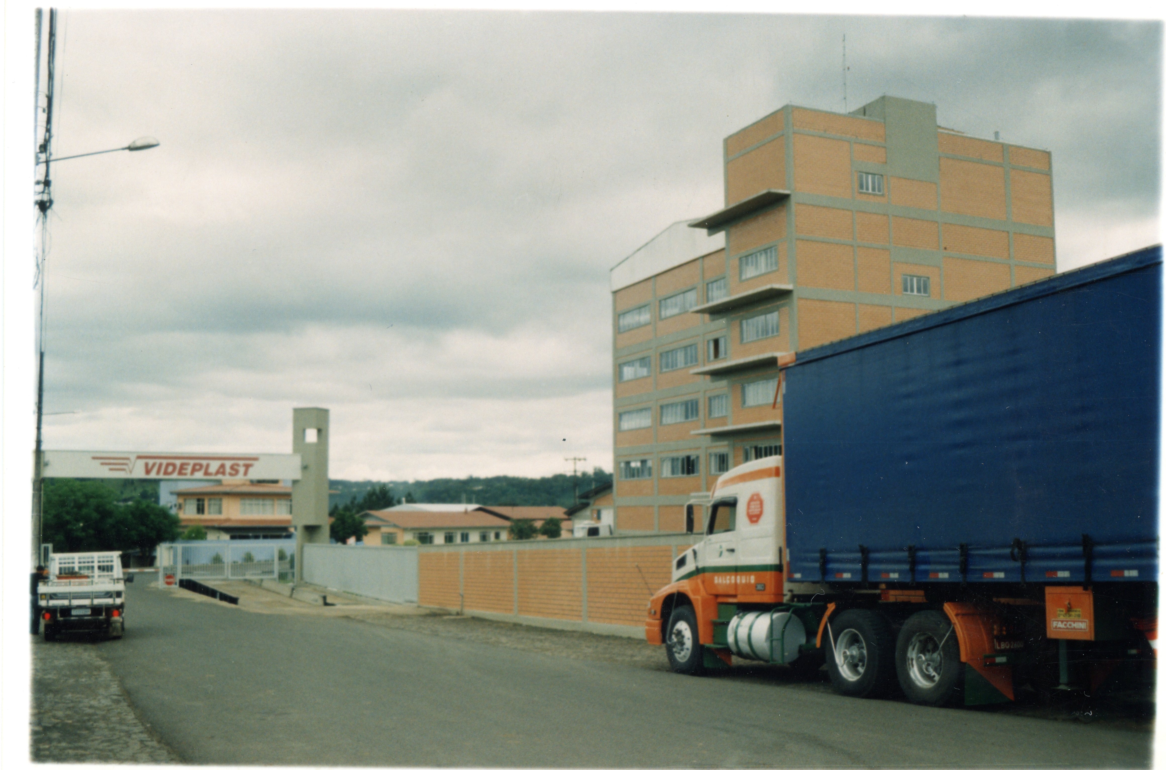A imagem mostra a entrada da empresa Videplast, com um caminhão estacionado em primeiro plano e um prédio industrial de tijolos aparentes ao fundo. O cenário reflete a paisagem urbana e industrial de Videira, Santa Catarina, provavelmente capturada entre o final da década de 1990 e início dos anos 2000.