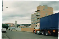 A imagem mostra a entrada da empresa Videplast, com um caminhão estacionado em primeiro plano e um prédio industrial de tijolos aparentes ao fundo. O cenário reflete a paisagem urbana e industrial de Videira, Santa Catarina, provavelmente capturada entre o final da década de 1990 e início dos anos 2000.