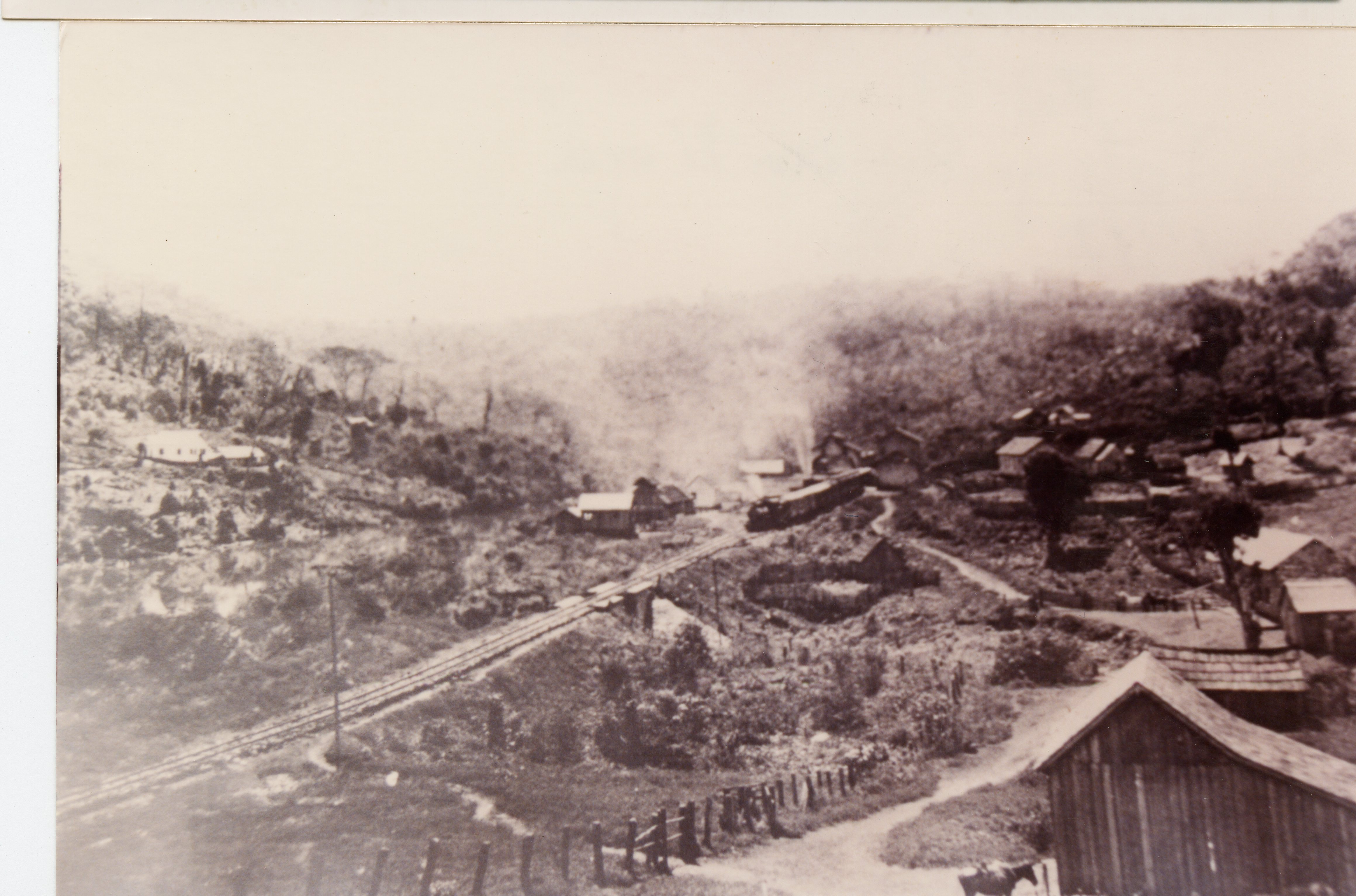 Fotografia histórica em preto e branco retratando um pequeno povoado rural brasileiro, possivelmente na região de Videira, com uma locomotiva a vapor cruzando uma ponte ferroviária. A paisagem é marcada por colinas, vegetação nativa e construções de madeira simples, sugerindo o período de expansão das ferrovias no início do século XX.