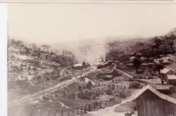 Fotografia histórica em preto e branco retratando um pequeno povoado rural brasileiro, possivelmente na região de Videira, com uma locomotiva a vapor cruzando uma ponte ferroviária. A paisagem é marcada por colinas, vegetação nativa e construções de madeira simples, sugerindo o período de expansão das ferrovias no início do século XX.