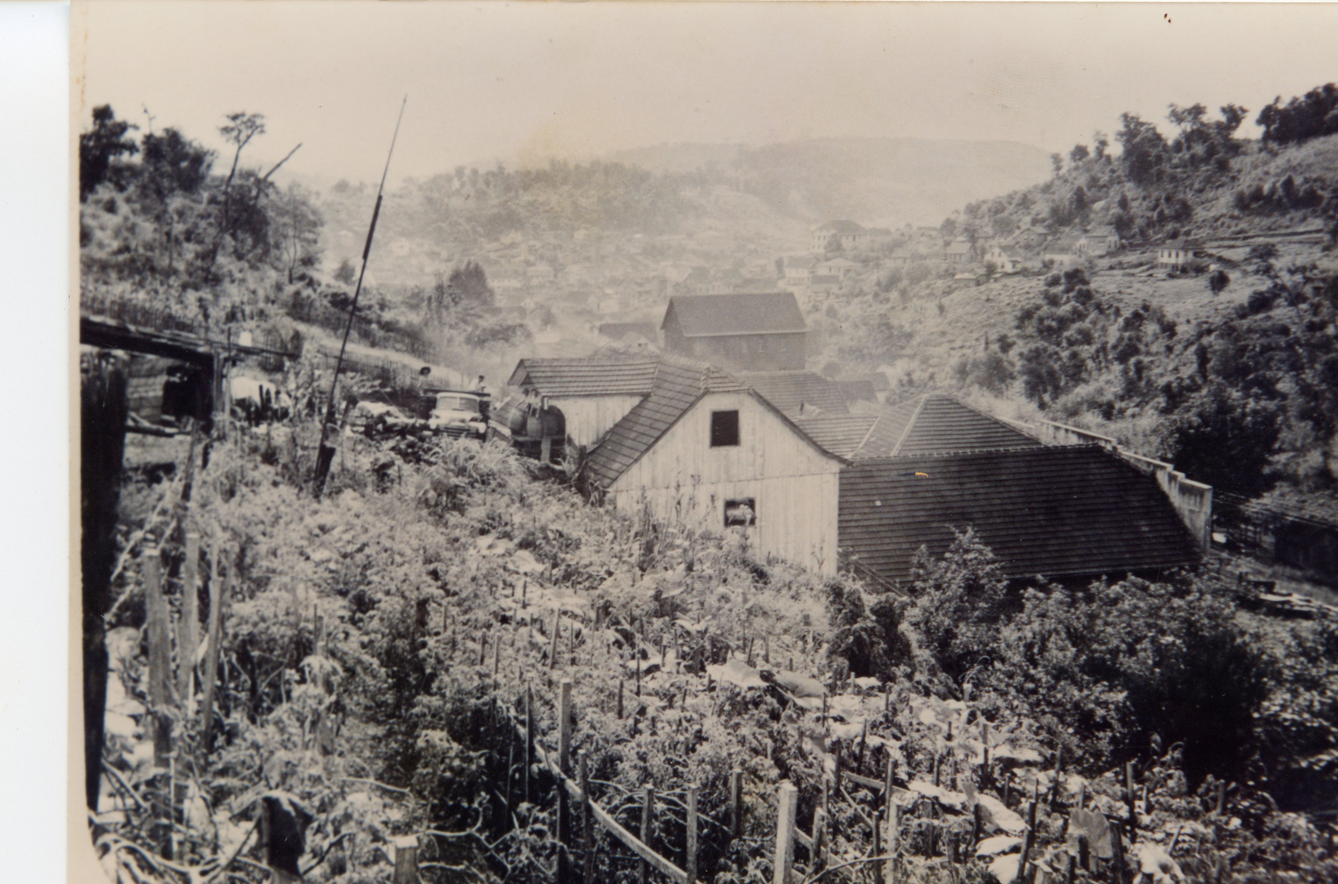 Fotografia em preto e branco de uma paisagem rural ou periurbana, possivelmente no interior do Brasil, exibindo casas de madeira com telhados de telha cerâmica e uma plantação em primeiro plano. A topografia montanhosa ao fundo sugere uma região de colonização, com um veículo antigo visível próximo às construções, indicando uma época entre as décadas de 1940 e 1960.