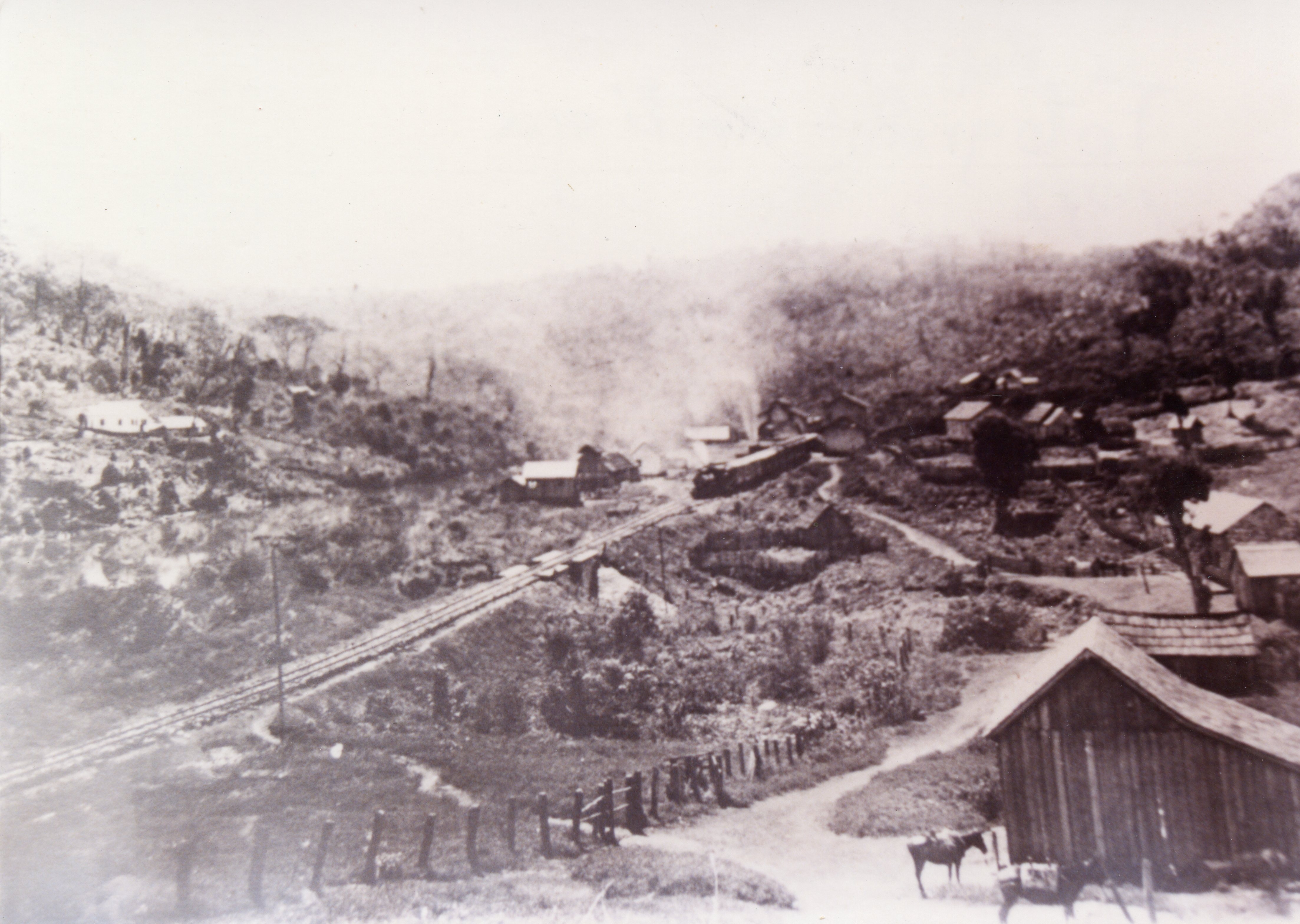 Fotografia histórica em preto e branco retratando um pequeno povoado rural atravessado por uma linha férrea, com uma locomotiva a vapor emitindo fumaça. O cenário apresenta construções de madeira simples, vegetação nativa e animais de carga, sugerindo o período de expansão ferroviária no interior do Brasil no início do século XX.