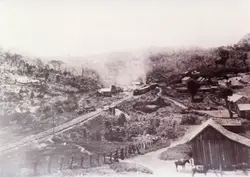 Fotografia histórica em preto e branco retratando um pequeno povoado rural atravessado por uma linha férrea, com uma locomotiva a vapor emitindo fumaça. O cenário apresenta construções de madeira simples, vegetação nativa e animais de carga, sugerindo o período de expansão ferroviária no interior do Brasil no início do século XX.