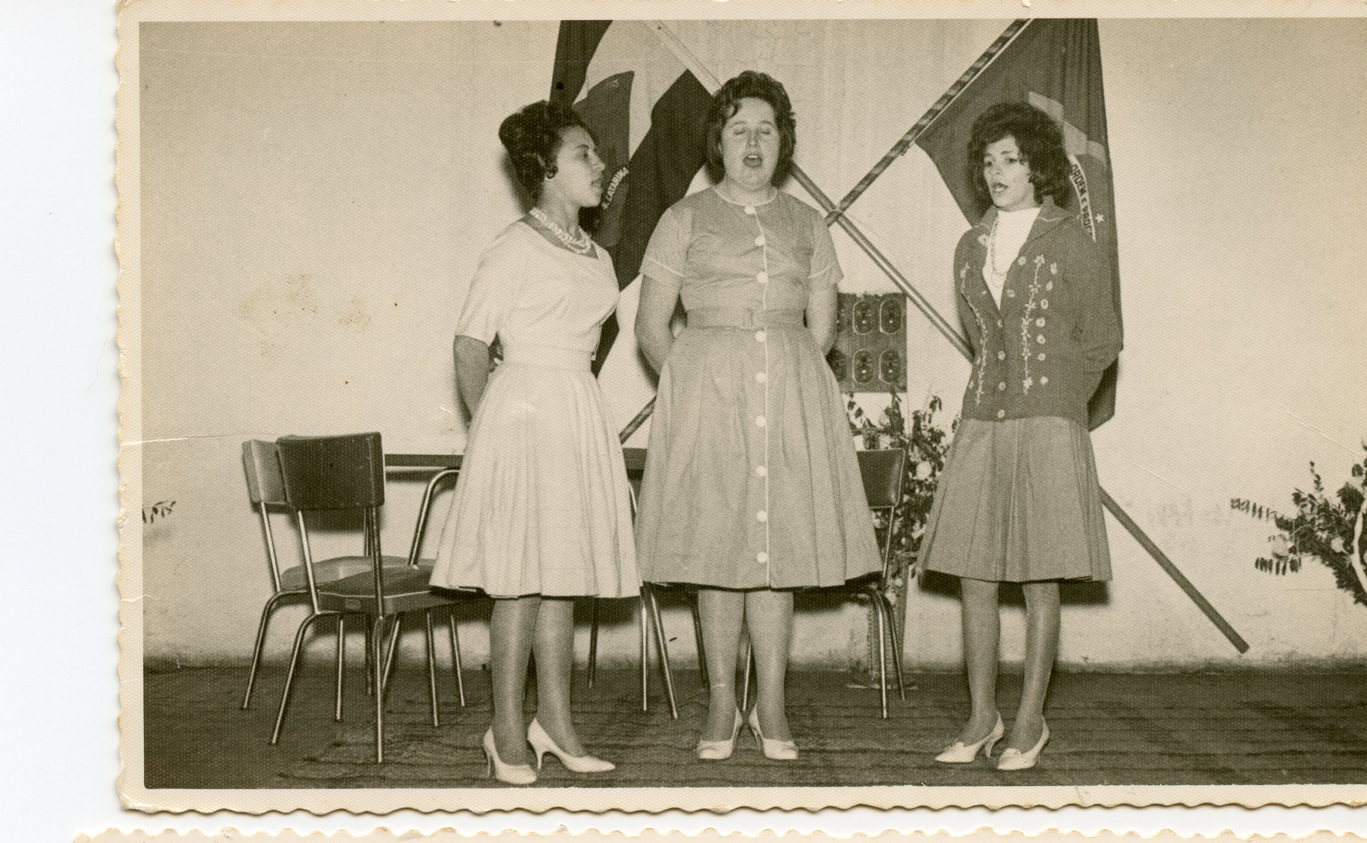 Três jovens mulheres aparecem em pé, cantando ou discursando, em um ambiente interno com bandeiras ao fundo, possivelmente em um evento cívico ou escolar. A vestimenta, com vestidos de corte clássico e penteados volumosos, sugere a moda típica da década de 1960 em um contexto brasileiro.