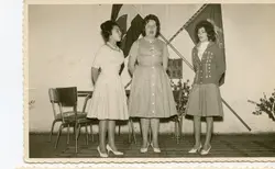 Três jovens mulheres aparecem em pé, cantando ou discursando, em um ambiente interno com bandeiras ao fundo, possivelmente em um evento cívico ou escolar. A vestimenta, com vestidos de corte clássico e penteados volumosos, sugere a moda típica da década de 1960 em um contexto brasileiro.
