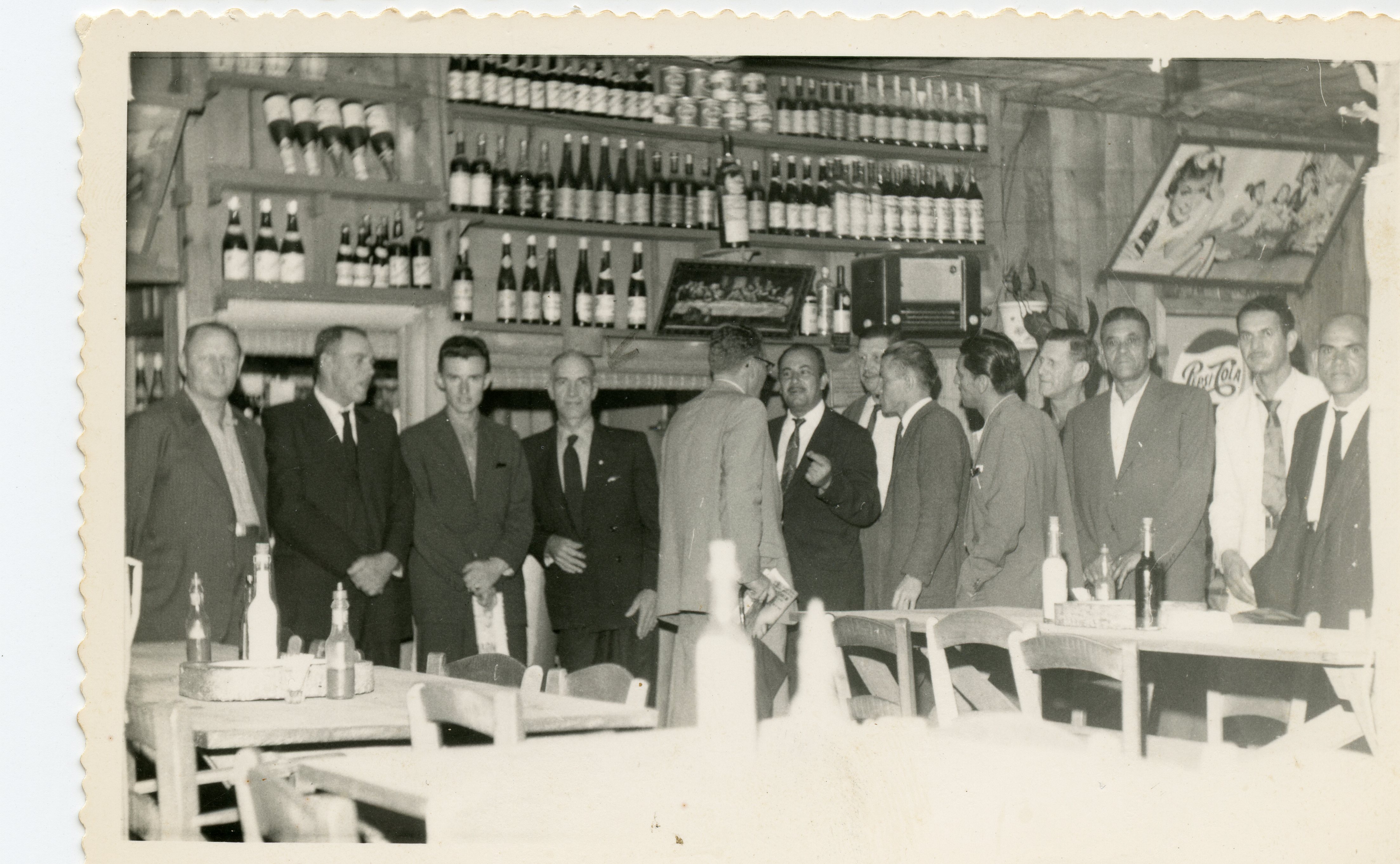 Fotografia em preto e branco de um grupo de homens reunidos em um estabelecimento comercial, possivelmente um bar ou restaurante, com prateleiras repletas de garrafas ao fundo. A cena sugere um encontro social ou de negócios típico de meados do século XX, com os homens vestindo ternos formais e um cartaz da Pepsi-Cola visível na parede.