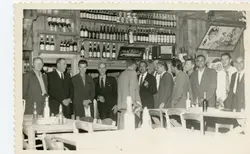 Fotografia em preto e branco de um grupo de homens reunidos em um estabelecimento comercial, possivelmente um bar ou restaurante, com prateleiras repletas de garrafas ao fundo. A cena sugere um encontro social ou de negócios típico de meados do século XX, com os homens vestindo ternos formais e um cartaz da Pepsi-Cola visível na parede.