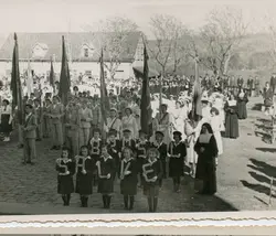 Fotografia em preto e branco de um desfile cívico ou escolar, possivelmente em uma cidade do interior do Brasil, com crianças segurando letras que formam a palavra 'BRASIL'. O cenário inclui uma construção de arquitetura simples ao fundo e a presença de religiosas, sugerindo um evento de uma escola católica ou instituição comunitária em meados do século XX.