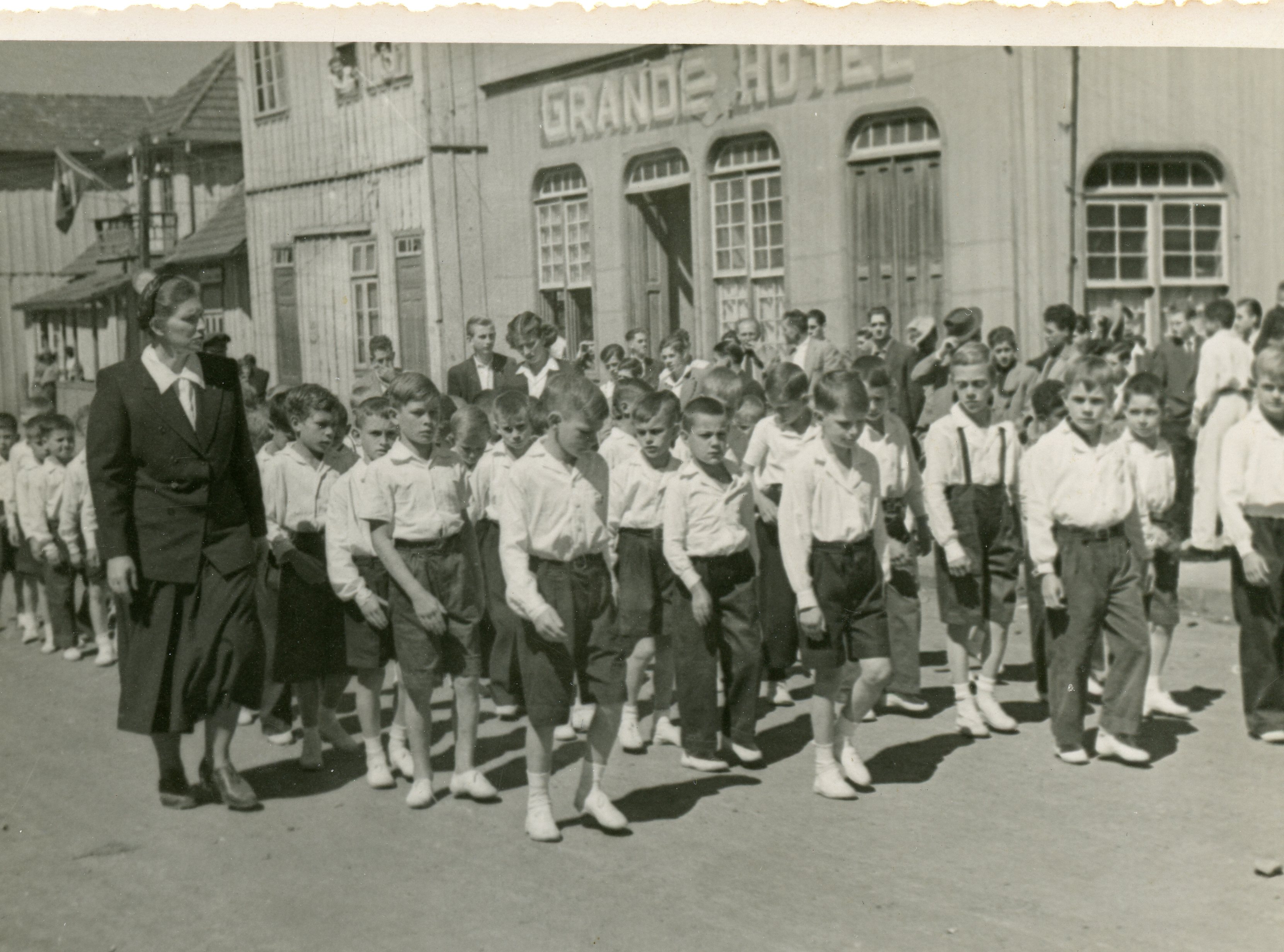 Uma fotografia em preto e branco que retrata um desfile ou procissão escolar em frente ao 'Grande Hotel', com uma professora liderando um grupo de meninos uniformizados. A arquitetura de madeira e o estilo das vestimentas sugerem uma pequena cidade brasileira em meados do século XX, possivelmente durante um evento cívico ou religioso.