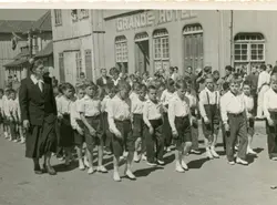 Uma fotografia em preto e branco que retrata um desfile ou procissão escolar em frente ao 'Grande Hotel', com uma professora liderando um grupo de meninos uniformizados. A arquitetura de madeira e o estilo das vestimentas sugerem uma pequena cidade brasileira em meados do século XX, possivelmente durante um evento cívico ou religioso.