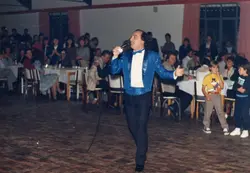 Uma fotografia de um evento social em um salão, onde um cantor vestido com um paletó azul brilhante se apresenta ao microfone. O público está sentado em mesas dispostas ao redor, sugerindo uma festa ou jantar comemorativo típico de meados da década de 1990.
