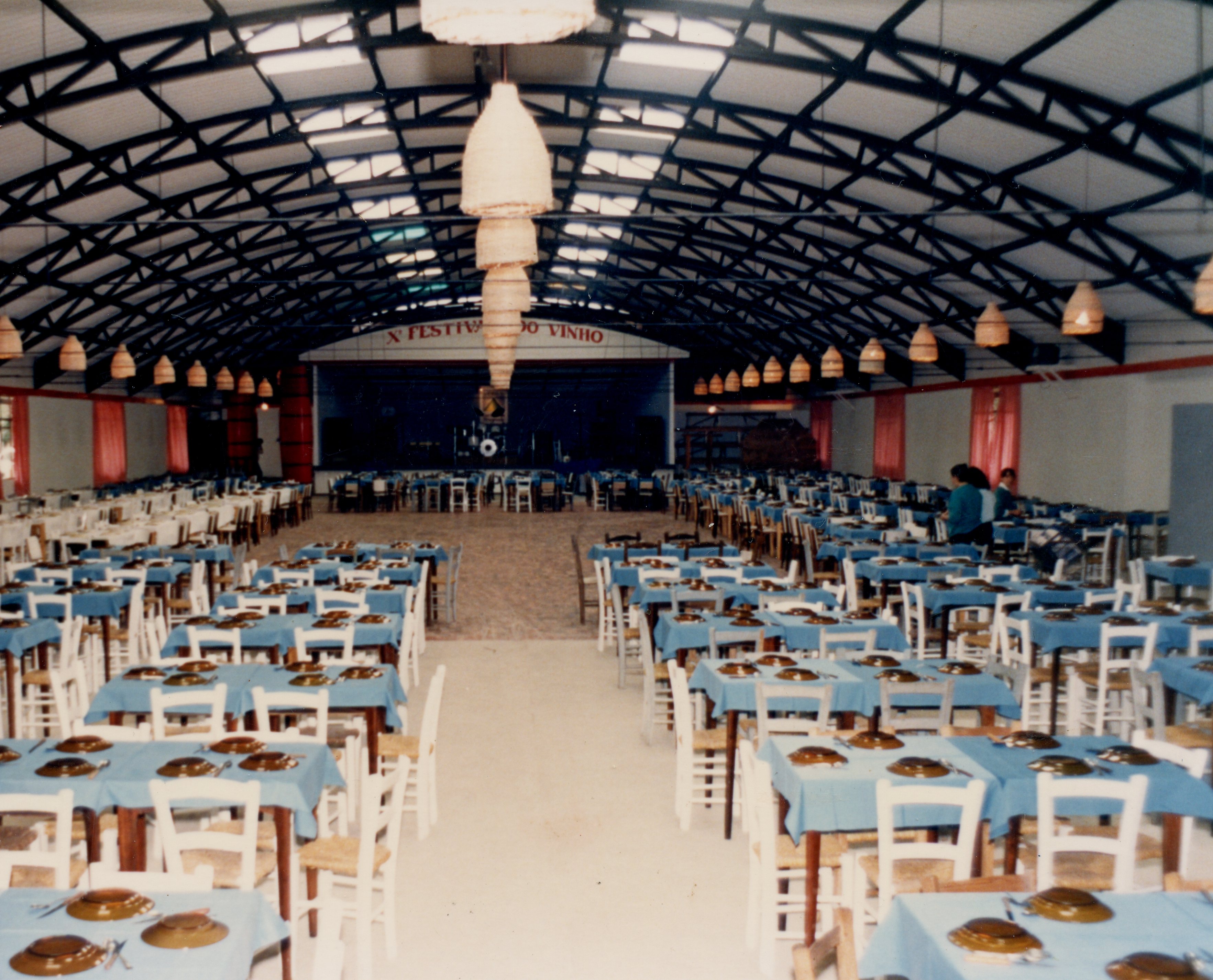 Uma fotografia de um amplo salão de eventos preparado para o 'X Festival do Vinho', com diversas mesas dispostas com toalhas azuis e cadeiras de madeira. O ambiente possui uma estrutura de teto metálico arqueado e iluminação pendente, sugerindo um evento comunitário ou festivo típico de cidades com tradição vitivinícola como Videira.