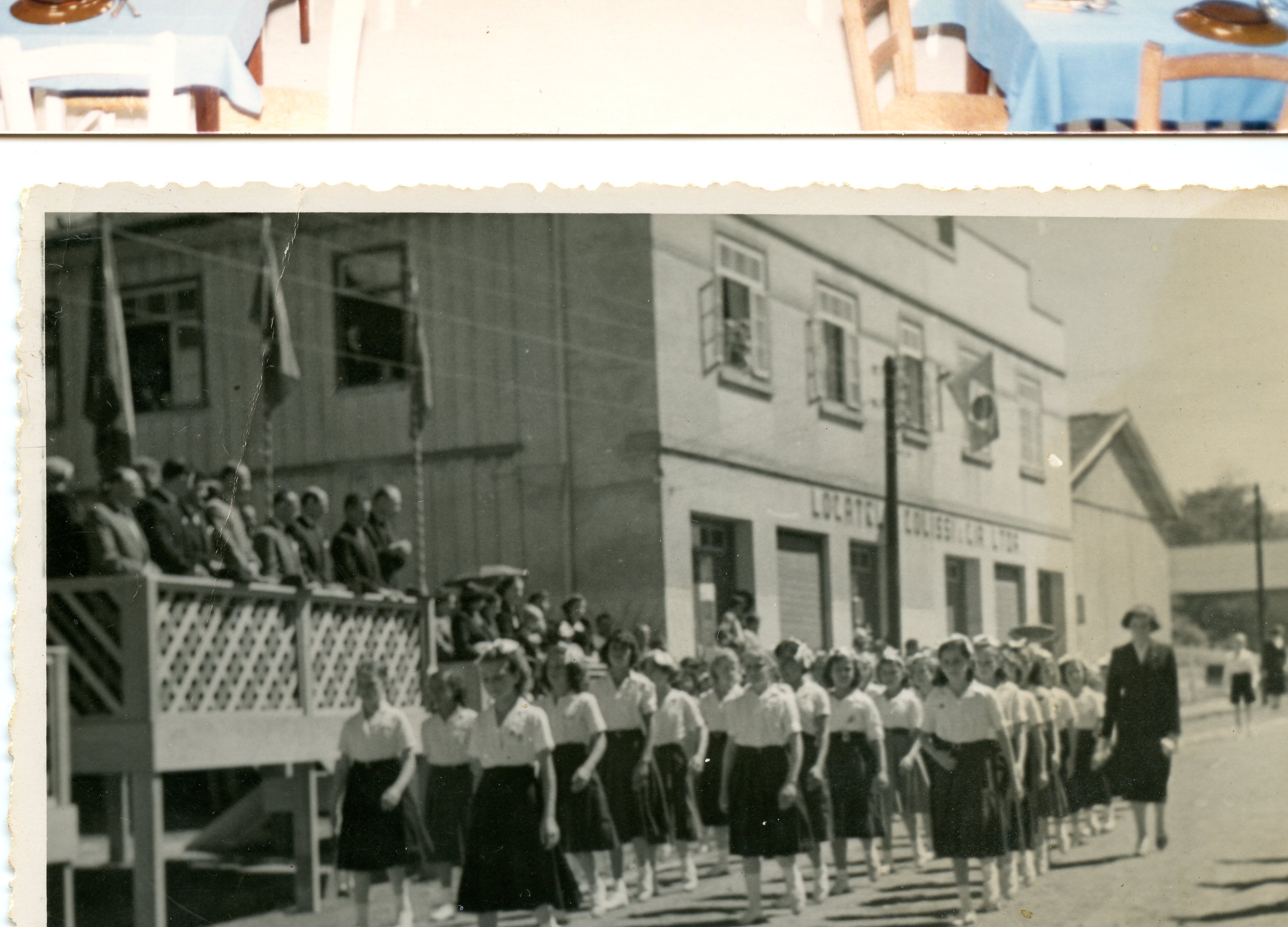 Uma fotografia em preto e branco que retrata um desfile cívico ou escolar em frente a um edifício comercial com a inscrição 'Locatelli Colissi & Cia Ltda'. O grupo de jovens, vestindo uniformes escolares, caminha pela rua enquanto autoridades observam de um palanque de madeira, sugerindo um evento comunitário em uma cidade brasileira de médio porte em meados do século XX.