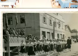 Uma fotografia em preto e branco que retrata um desfile cívico ou escolar em frente a um edifício comercial com a inscrição 'Locatelli Colissi & Cia Ltda'. O grupo de jovens, vestindo uniformes escolares, caminha pela rua enquanto autoridades observam de um palanque de madeira, sugerindo um evento comunitário em uma cidade brasileira de médio porte em meados do século XX.