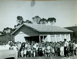 Uma fotografia em preto e branco que retrata um grande grupo de pessoas, incluindo muitas crianças, reunidas em frente a uma construção de madeira com telhado de telhas, possivelmente uma escola rural ou capela. A cena sugere um evento comunitário ou escolar em uma região de colonização, com vestimentas que indicam um período entre as décadas de 1950 e 1960.