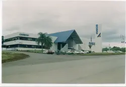 A imagem mostra a fachada de uma concessionária Carboni Iveco, caracterizada por uma arquitetura moderna com telhado em formato de 'A' e um totem vertical com a marca. O cenário sugere um ambiente comercial em uma região brasileira, possivelmente em Videira, com veículos estacionados e uma motocicleta em movimento no pátio.
