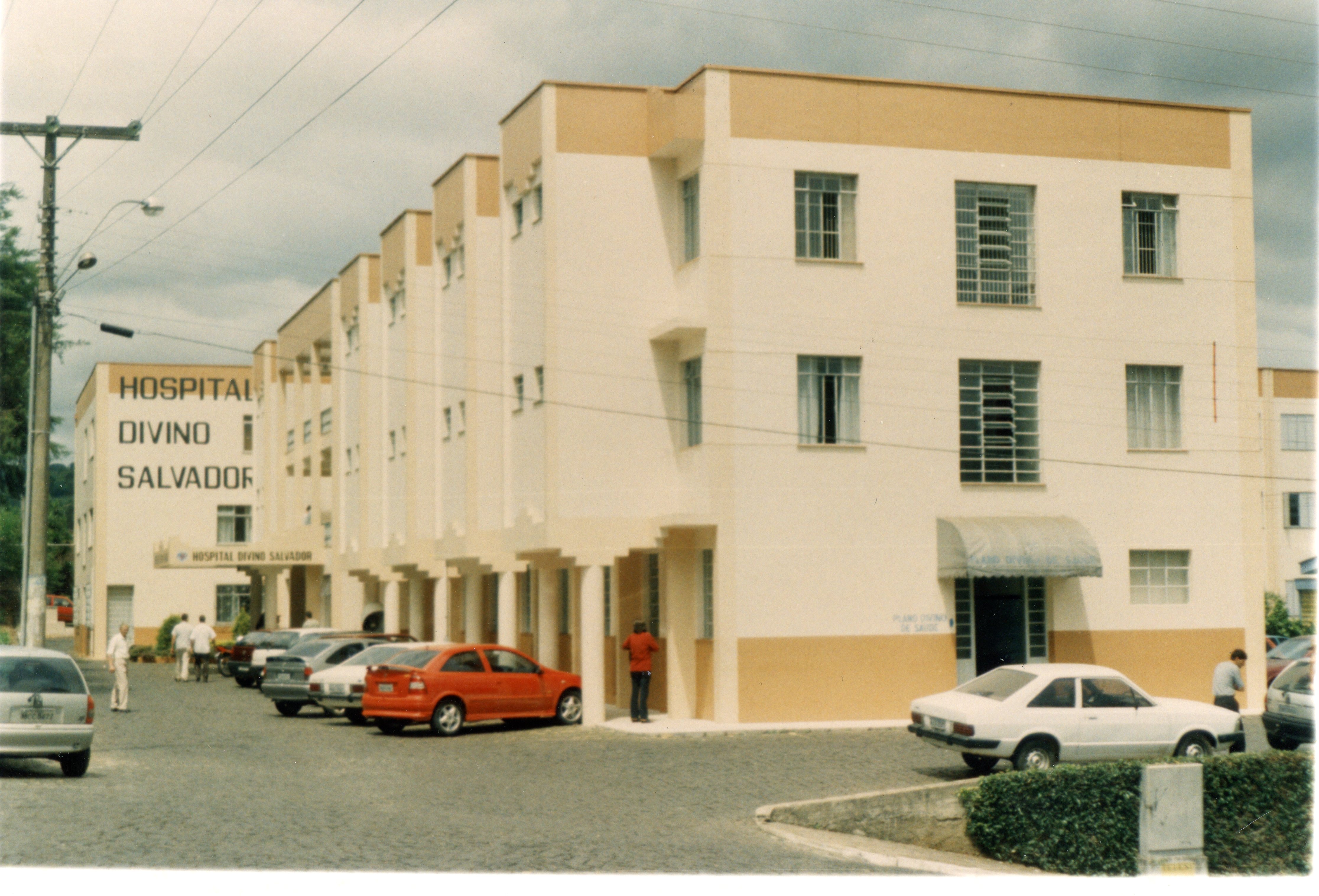 Fotografia externa do Hospital Divino Salvador, exibindo sua arquitetura institucional com tons de bege e branco. A imagem mostra o pátio frontal com diversos veículos estacionados e algumas pessoas circulando, sugerindo um registro cotidiano de uma cidade brasileira, possivelmente Videira, entre o final do século XX e início do XXI.