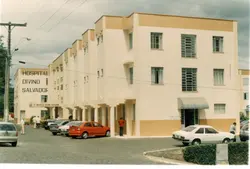 Fotografia externa do Hospital Divino Salvador, exibindo sua arquitetura institucional com tons de bege e branco. A imagem mostra o pátio frontal com diversos veículos estacionados e algumas pessoas circulando, sugerindo um registro cotidiano de uma cidade brasileira, possivelmente Videira, entre o final do século XX e início do XXI.