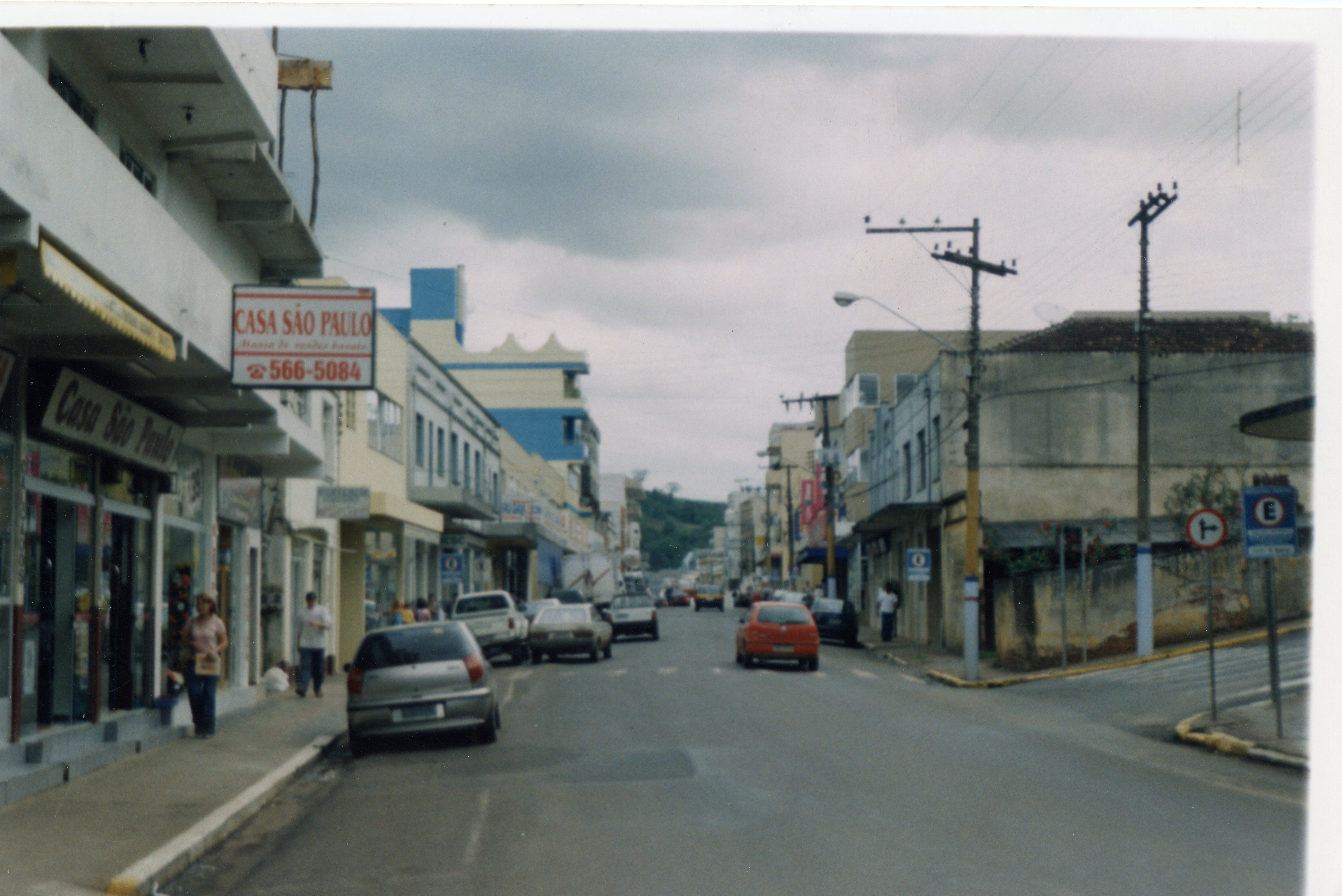 Uma vista de rua urbana em uma cidade brasileira, provavelmente no final da década de 1990 ou início dos anos 2000, caracterizada por edifícios comerciais de arquitetura simples e veículos populares da época. A imagem destaca a fachada da 'Casa São Paulo' e a movimentação cotidiana de pedestres e carros em um dia nublado.
