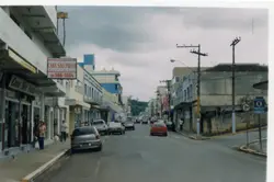 Uma vista de rua urbana em uma cidade brasileira, provavelmente no final da década de 1990 ou início dos anos 2000, caracterizada por edifícios comerciais de arquitetura simples e veículos populares da época. A imagem destaca a fachada da 'Casa São Paulo' e a movimentação cotidiana de pedestres e carros em um dia nublado.