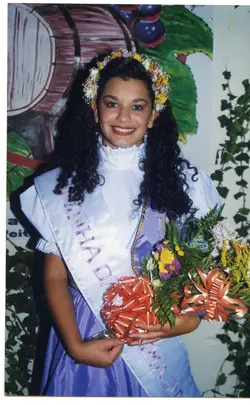 Uma jovem mulher posa para um retrato vestindo uma faixa de concurso de beleza, possivelmente uma rainha de festividade local, como as tradicionais festas da uva. A imagem apresenta elementos típicos dos anos 90, como o estilo do cabelo e a vestimenta, com um fundo decorativo pintado que remete a uma temática rural ou vitivinícola.