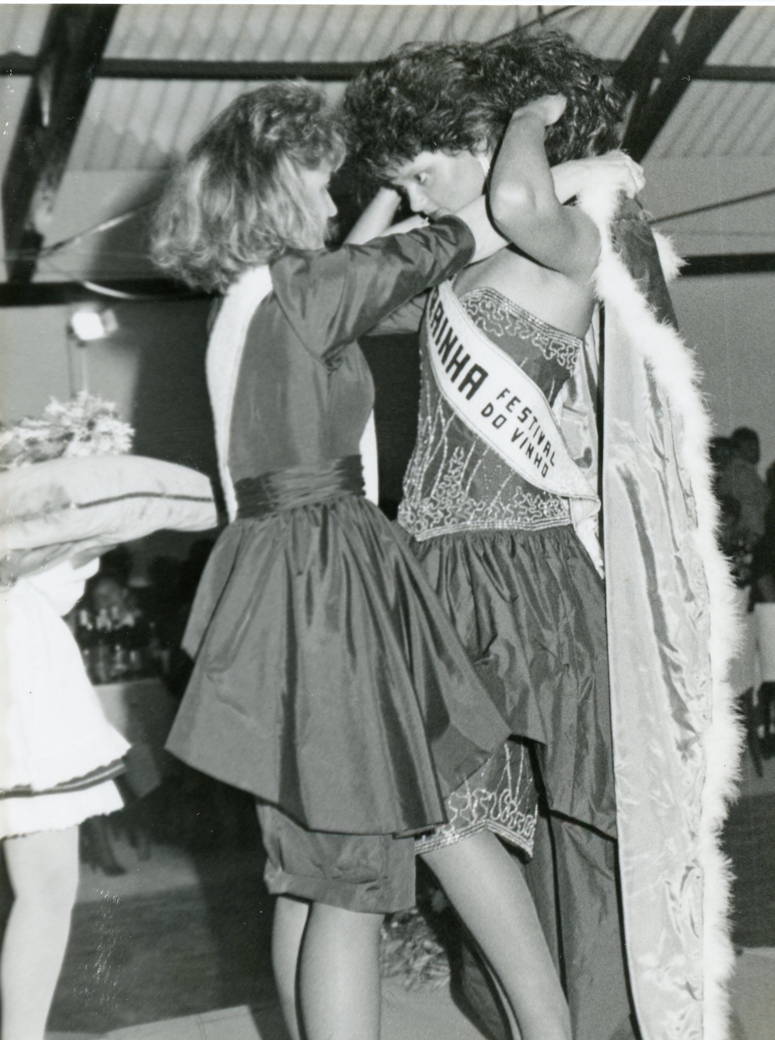 Fotografia em preto e branco registrando o momento da coroação de uma rainha em um concurso de beleza, possivelmente o Festival do Vinho. A imagem destaca o uso de faixas, vestidos de gala e uma capa com detalhes em pele, típicos de eventos sociais e festividades regionais brasileiras das décadas de 1980 ou 1990.