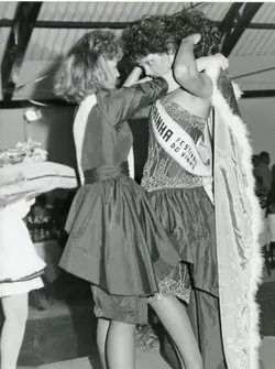 Fotografia em preto e branco registrando o momento da coroação de uma rainha em um concurso de beleza, possivelmente o Festival do Vinho. A imagem destaca o uso de faixas, vestidos de gala e uma capa com detalhes em pele, típicos de eventos sociais e festividades regionais brasileiras das décadas de 1980 ou 1990.