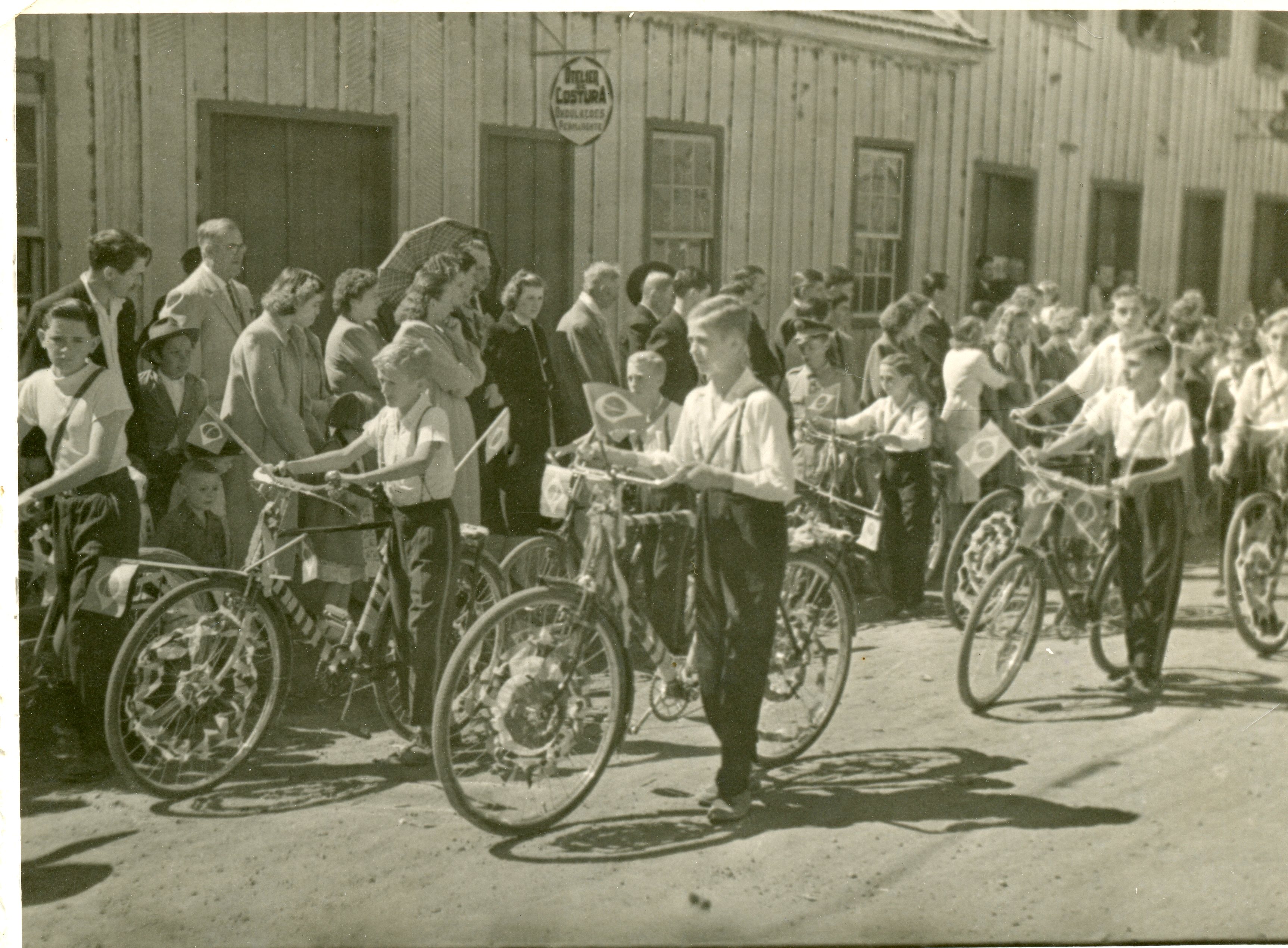 Uma fotografia em preto e branco que retrata um desfile cívico ou evento comemorativo em uma rua de terra, com crianças conduzindo bicicletas decoradas com bandeiras do Brasil. O cenário apresenta construções de madeira típicas do início da colonização, sugerindo uma pequena cidade brasileira em meados do século XX.