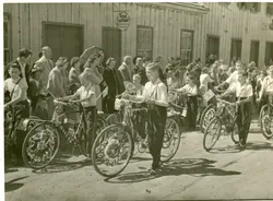 Uma fotografia em preto e branco que retrata um desfile cívico ou evento comemorativo em uma rua de terra, com crianças conduzindo bicicletas decoradas com bandeiras do Brasil. O cenário apresenta construções de madeira típicas do início da colonização, sugerindo uma pequena cidade brasileira em meados do século XX.