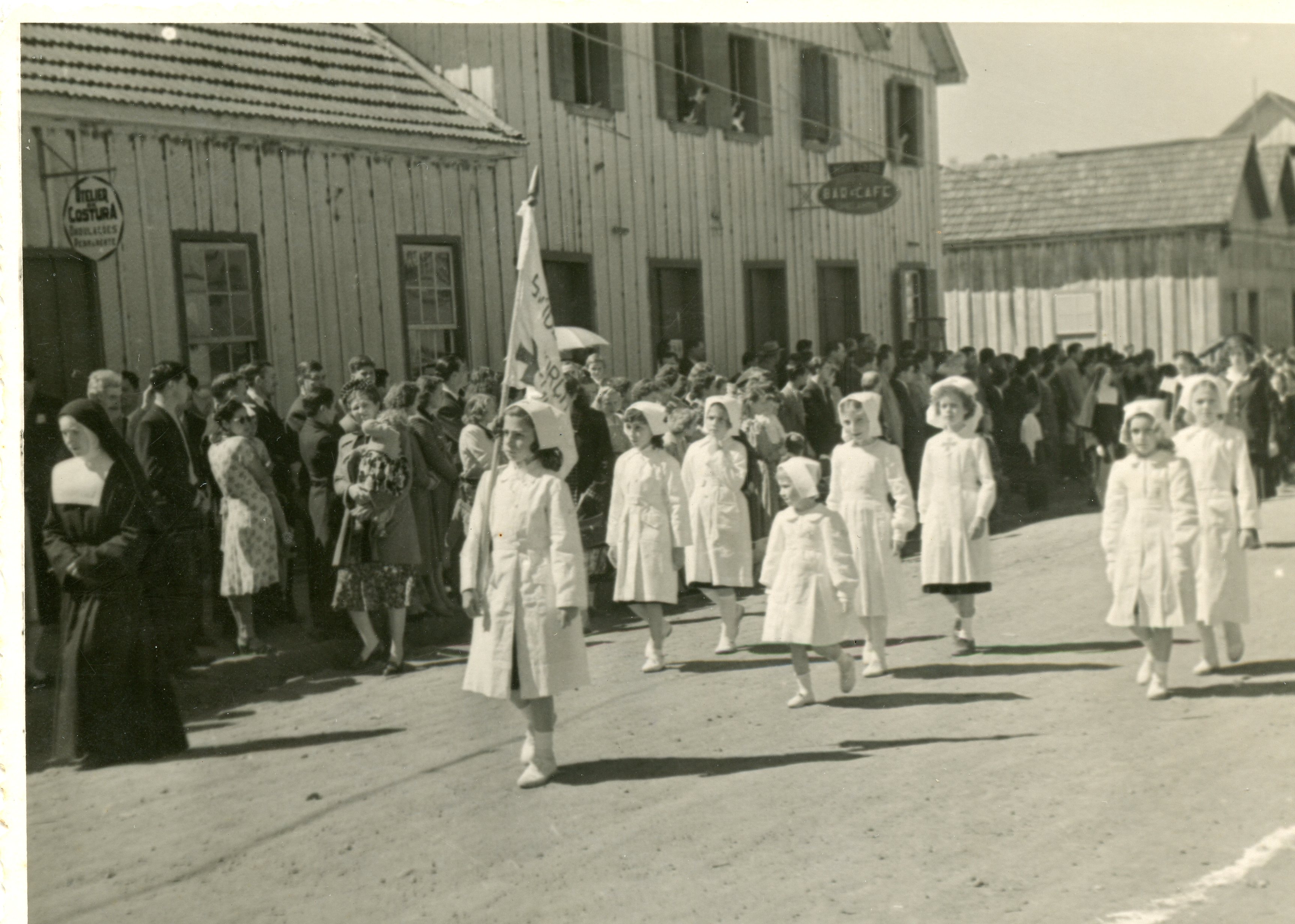 Uma fotografia em preto e branco que retrata um desfile ou procissão em uma rua de terra, com um grupo de meninas vestidas de enfermeiras caminhando à frente. Ao fundo, observam-se construções de madeira típicas da colonização sulista brasileira, incluindo um estabelecimento com placa de 'Bar-Café' e um 'Atelier de Costura', sugerindo um contexto de meados do século XX.