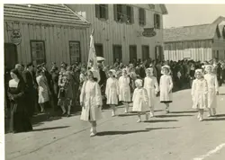 Uma fotografia em preto e branco que retrata um desfile ou procissão em uma rua de terra, com um grupo de meninas vestidas de enfermeiras caminhando à frente. Ao fundo, observam-se construções de madeira típicas da colonização sulista brasileira, incluindo um estabelecimento com placa de 'Bar-Café' e um 'Atelier de Costura', sugerindo um contexto de meados do século XX.