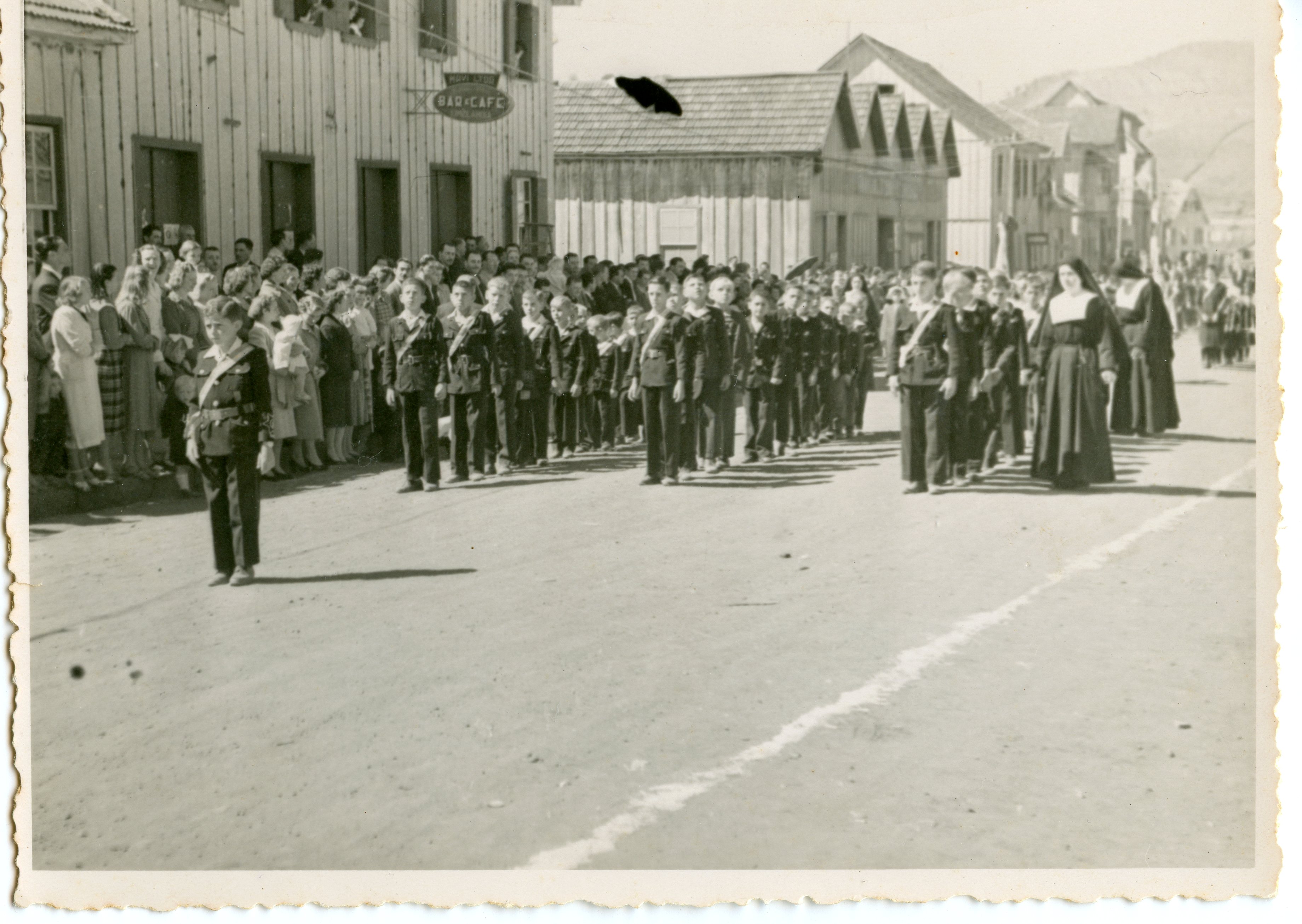 Fotografia histórica em preto e branco retratando um desfile cívico ou religioso em uma rua de terra, com crianças uniformizadas marchando e uma freira acompanhando o grupo. O cenário apresenta construções de madeira típicas da colonização sulista brasileira, sugerindo um ambiente urbano em desenvolvimento na primeira metade do século XX.