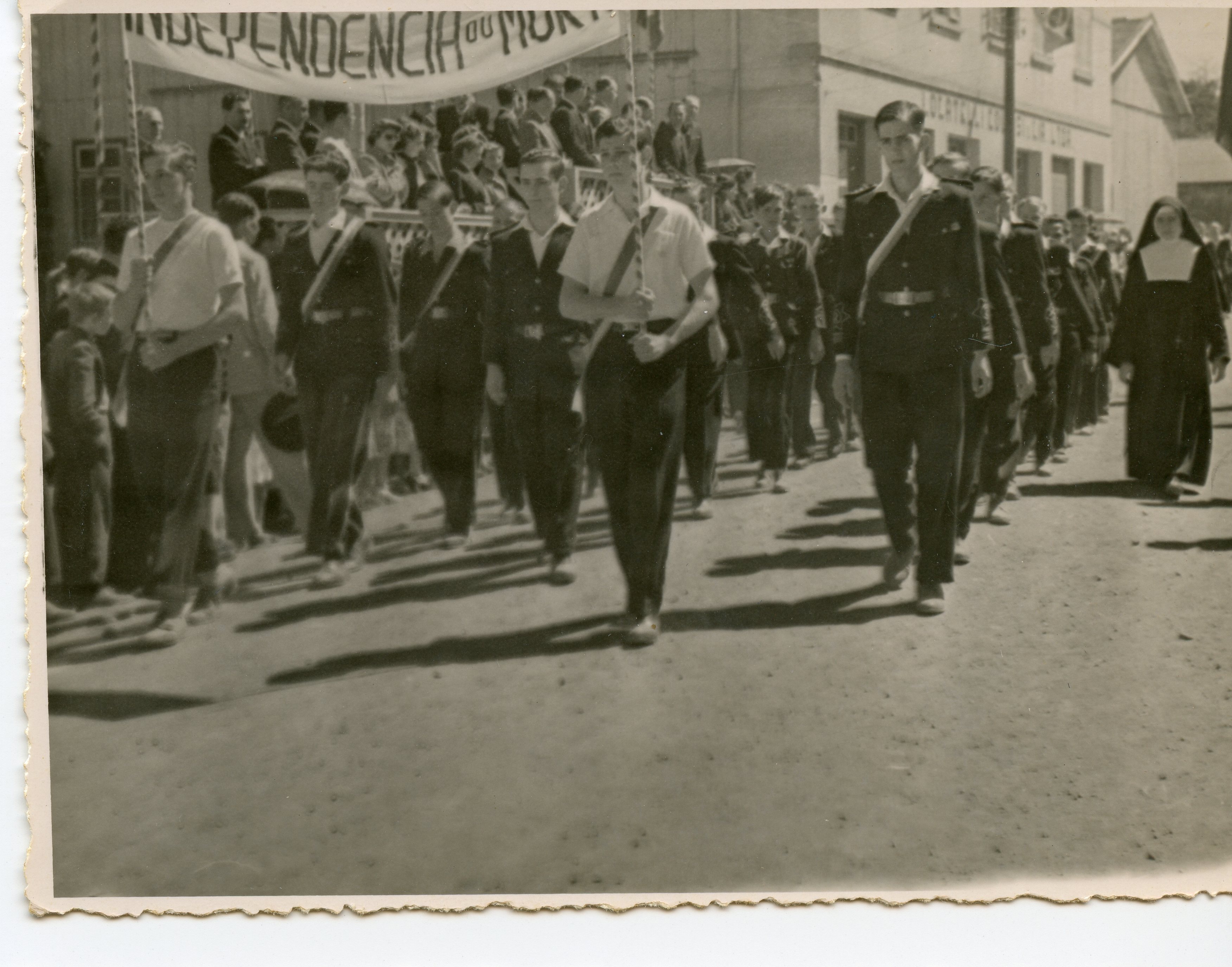 Fotografia em preto e branco de um desfile cívico, provavelmente em uma cidade brasileira, com jovens marchando e carregando uma faixa com os dizeres 'Independência ou Morte'. A cena inclui uma freira e estudantes uniformizados, sugerindo um evento escolar ou comemorativo de meados do século XX.