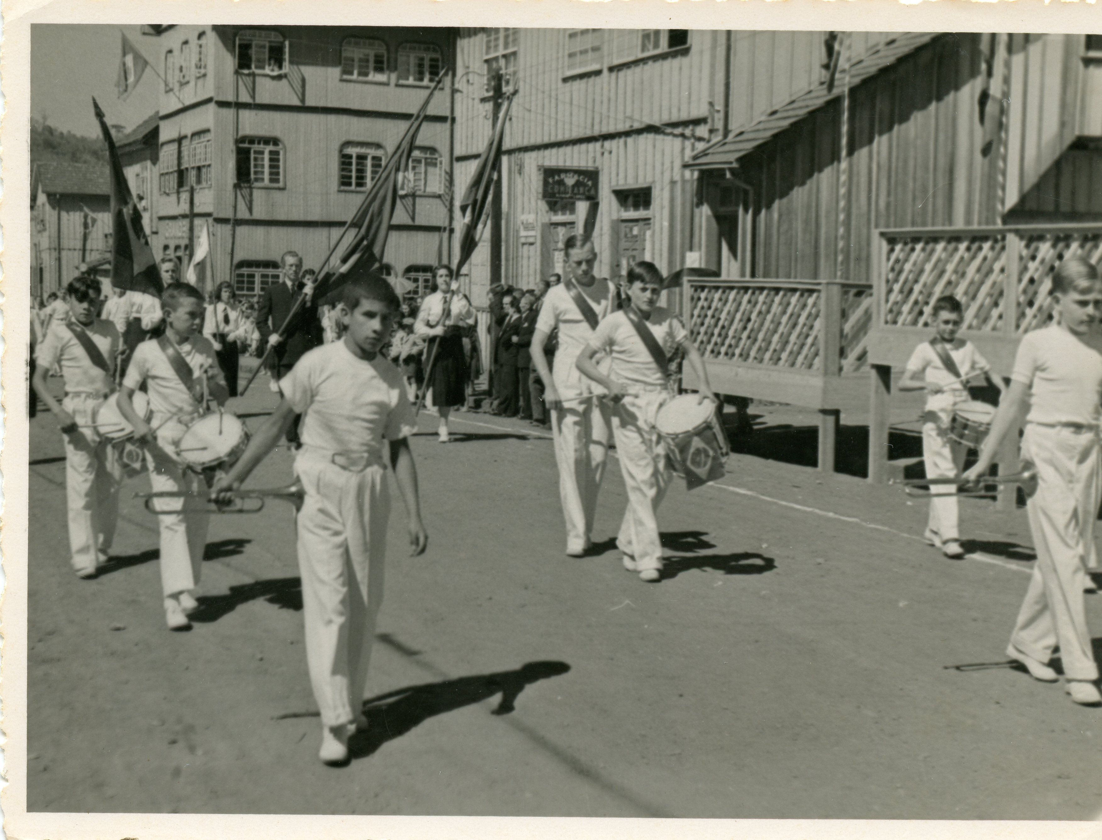 Fotografia em preto e branco de um desfile cívico ou escolar em uma pequena cidade brasileira, possivelmente na região sul. Jovens uniformizados com camisas brancas e faixas a tiracolo marcham tocando instrumentos de percussão e trompetes, em frente a construções de madeira típicas da colonização local.