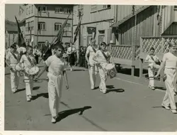 Fotografia em preto e branco de um desfile cívico ou escolar em uma pequena cidade brasileira, possivelmente na região sul. Jovens uniformizados com camisas brancas e faixas a tiracolo marcham tocando instrumentos de percussão e trompetes, em frente a construções de madeira típicas da colonização local.