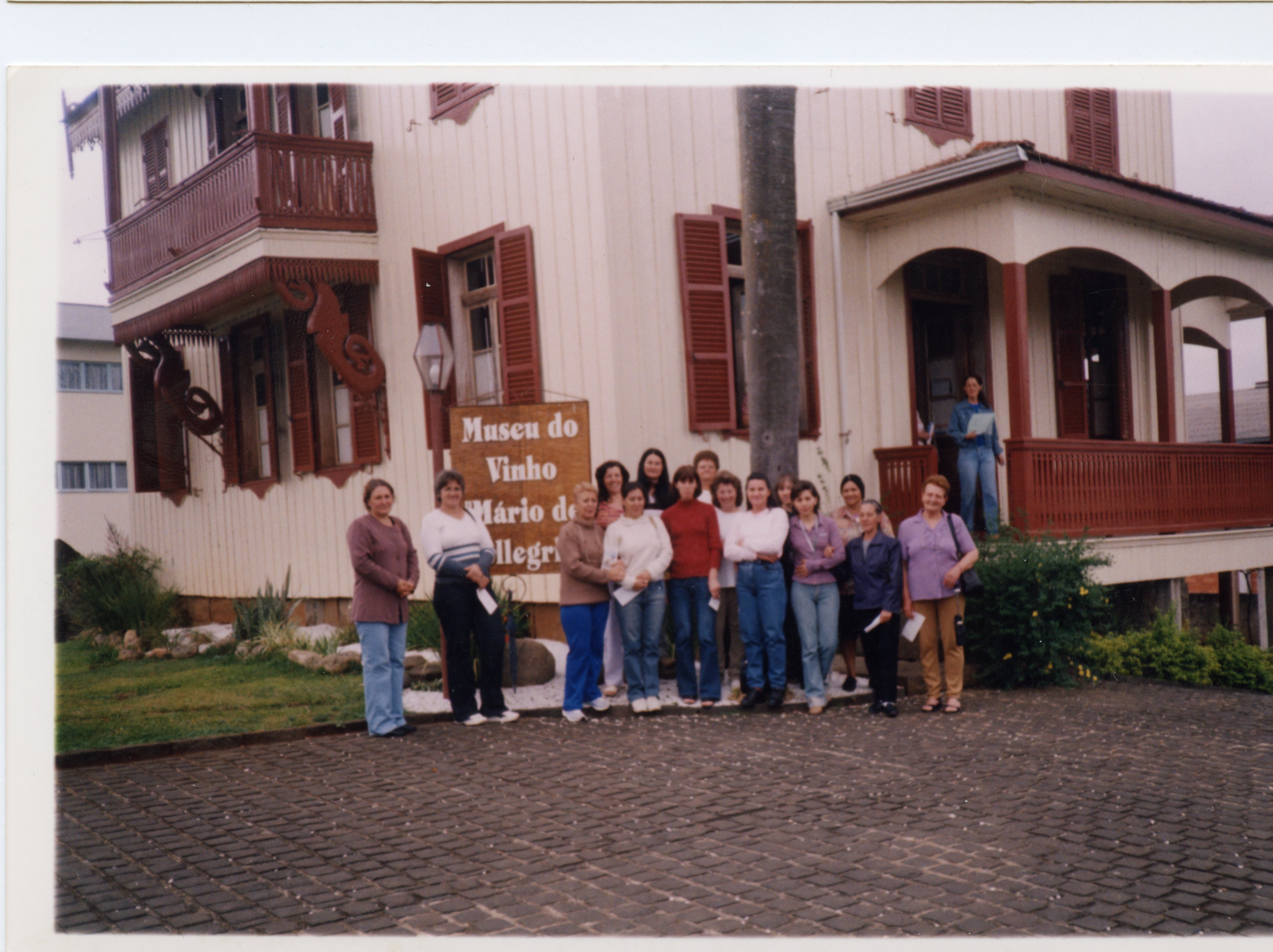 Uma fotografia de um grupo de mulheres posando em frente ao Museu do Vinho Mário de Pellegrin, em Videira, Santa Catarina. A arquitetura de madeira com detalhes em vermelho e o estilo das roupas sugerem uma visita turística ou cultural ocorrida provavelmente no final do século XX ou início do XXI.