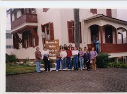 Uma fotografia de um grupo de mulheres posando em frente ao Museu do Vinho Mário de Pellegrin, em Videira, Santa Catarina. A arquitetura de madeira com detalhes em vermelho e o estilo das roupas sugerem uma visita turística ou cultural ocorrida provavelmente no final do século XX ou início do XXI.