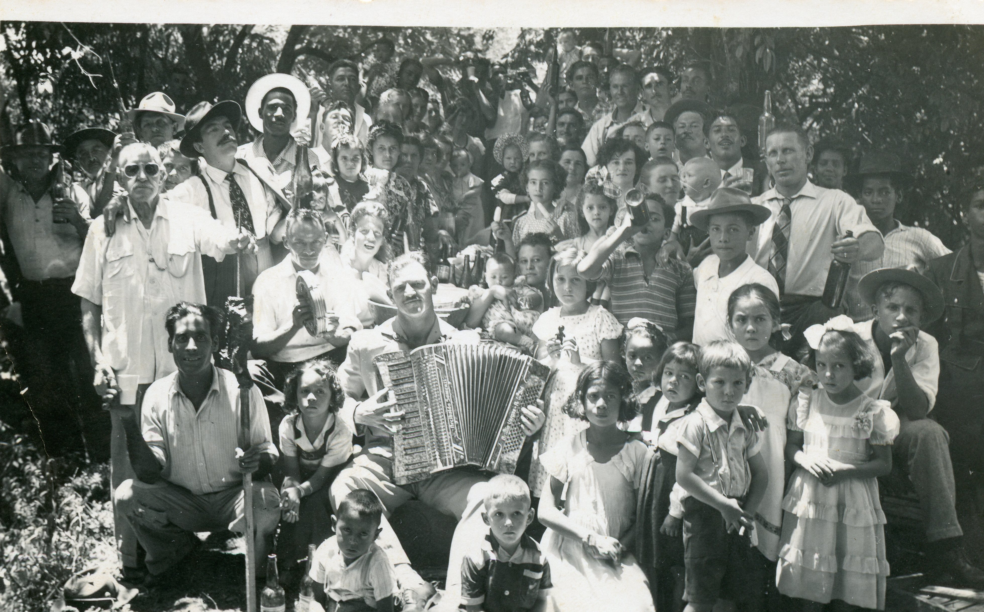 Uma fotografia em preto e branco que retrata uma grande reunião social ao ar livre, possivelmente um piquenique ou celebração comunitária em uma área rural brasileira. O grupo é composto por homens, mulheres e muitas crianças, com um homem ao centro tocando acordeão, sugerindo um ambiente festivo e de confraternização típica de meados do século XX.