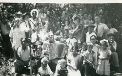 Uma fotografia em preto e branco que retrata uma grande reunião social ao ar livre, possivelmente um piquenique ou celebração comunitária em uma área rural brasileira. O grupo é composto por homens, mulheres e muitas crianças, com um homem ao centro tocando acordeão, sugerindo um ambiente festivo e de confraternização típica de meados do século XX.