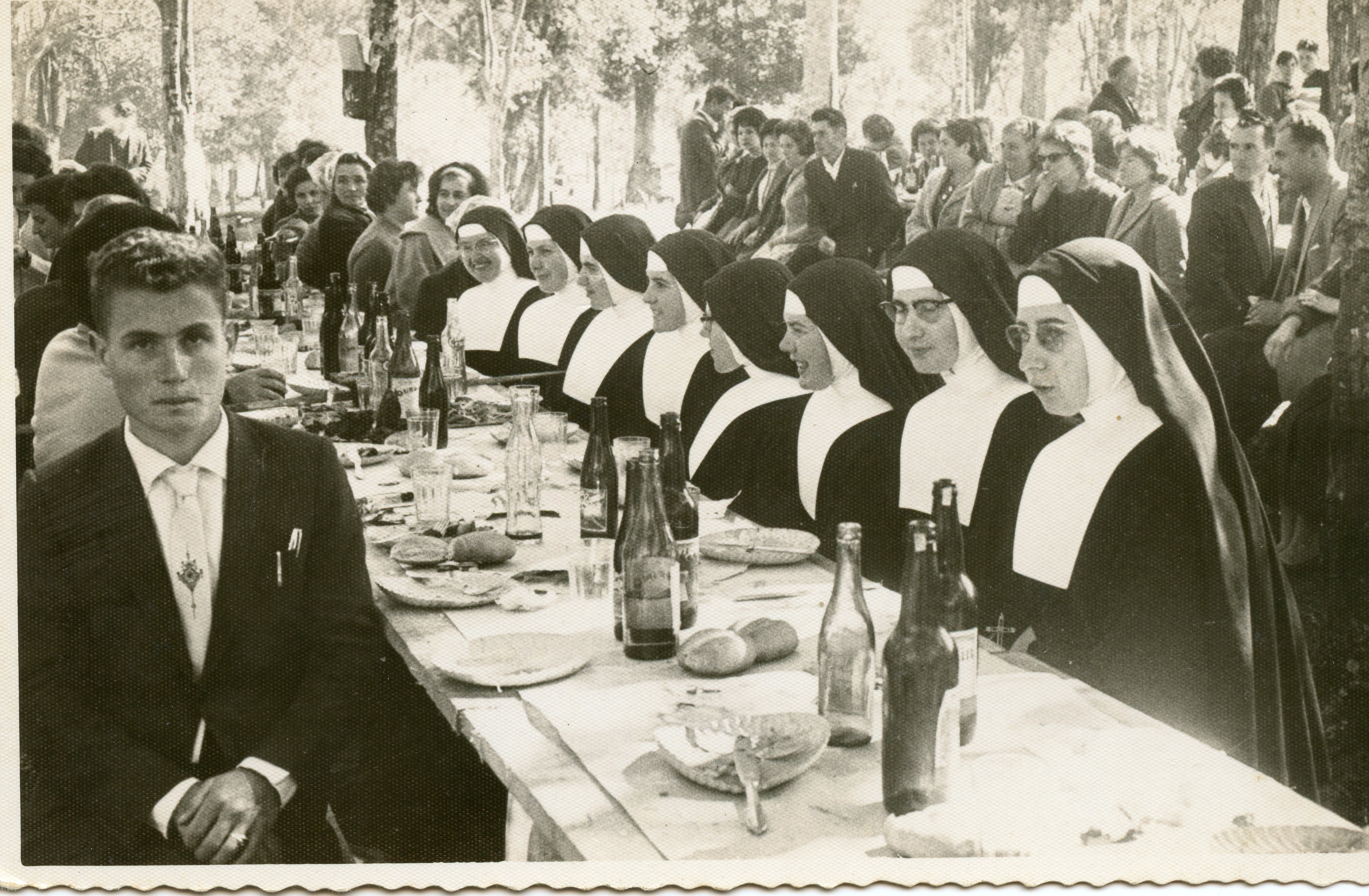Uma fotografia em preto e branco que retrata um banquete ao ar livre, possivelmente um evento comunitário ou religioso, com um grupo de freiras sentadas à mesa ao lado de um homem em primeiro plano. A cena sugere um contexto de meados do século XX, com vestimentas formais e hábitos religiosos tradicionais, refletindo um momento de confraternização em um ambiente arborizado.